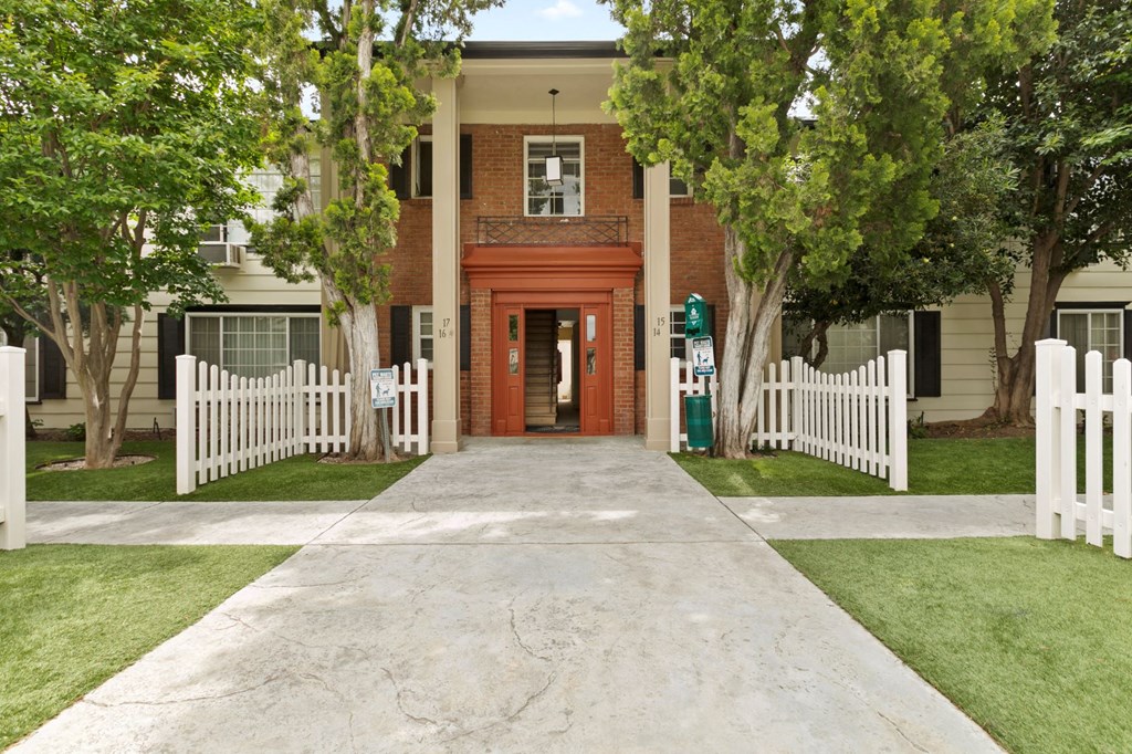 Apartments for Rent in Van Nuys CA - Colonial Manor - Exterior View Of Apartment Complex Entrance Featuring Stone Walkway, Lush Green Grass and Trees, And White Picket Fence