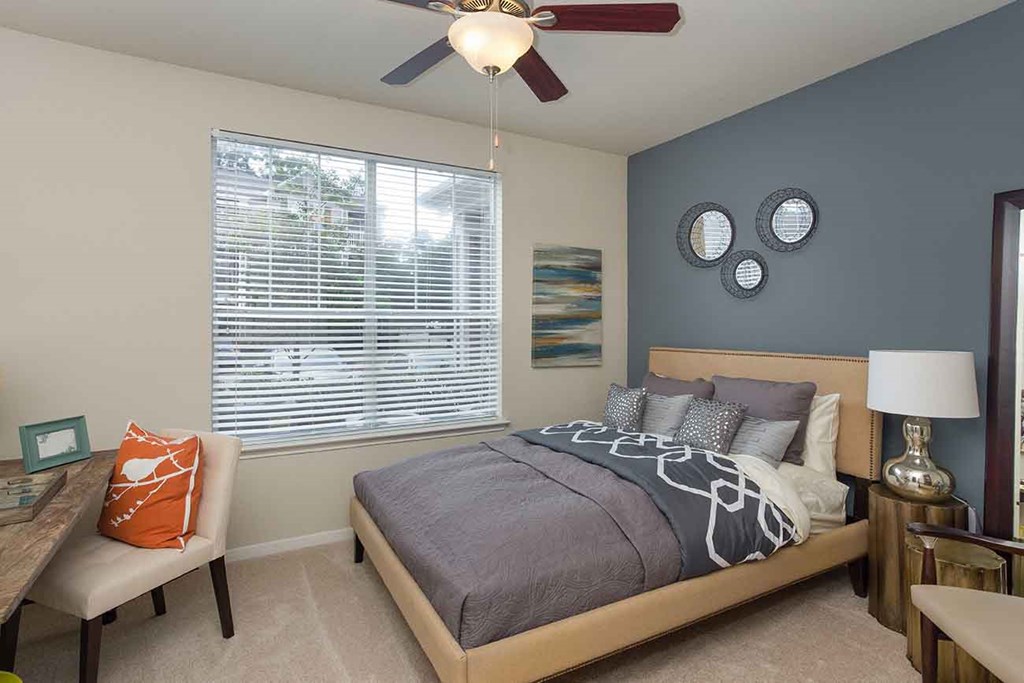 Bedroom Apartments for Rent in The Woodlands TX - Whispering Pines Ranch - Furnished Bedroom With Comfortable Bed, a Ceiling Fan, and a Desk With a Chair on Top of Carpet Flooring
