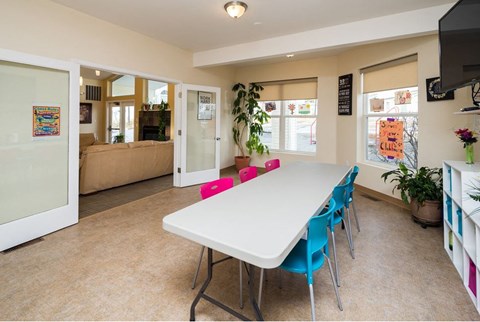 A white dining table with blue chairs is in the center of a room with a glass door leading to a living room.