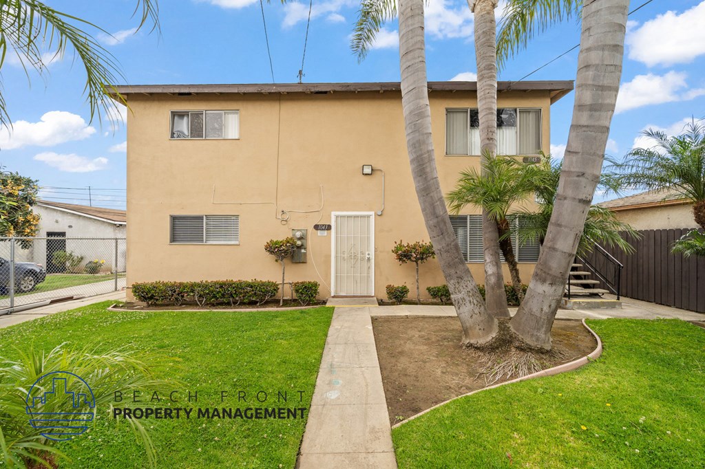 A house with a tan exterior and a white door is surrounded by a green lawn and palm trees.
