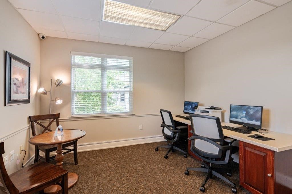 a room with a desk with a computer and chairs and a window