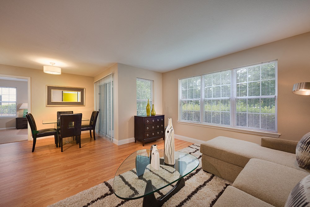 Furnished apartment living room showing abundant windows, balcony, and dining area in the background.