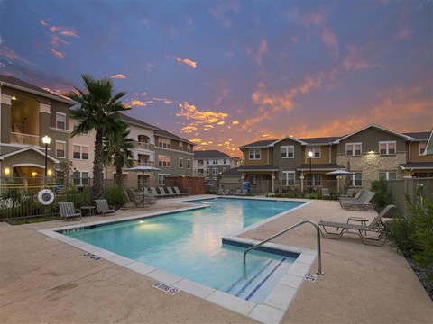 A swimming pool surrounded by lounge chairs and apartment buildings under a twilight sky.