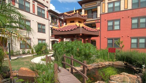 Beautifully landscaped community courtyard featuring a charming footbridge, lush greenery, and inviting outdoor spaces surrounded by apartment homes.