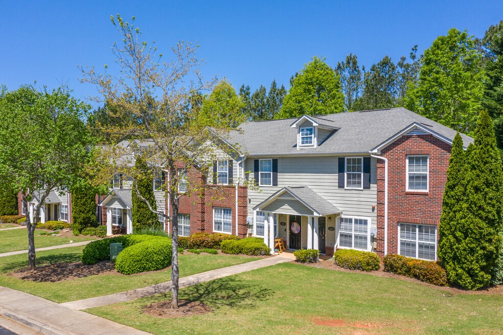 Charming two-story apartment building with a mix of brick and siding exterior, surrounded by lush greenery, manicured bushes, and mature trees. A concrete walkway leads to the covered entrance, and large windows enhance the inviting curb appeal