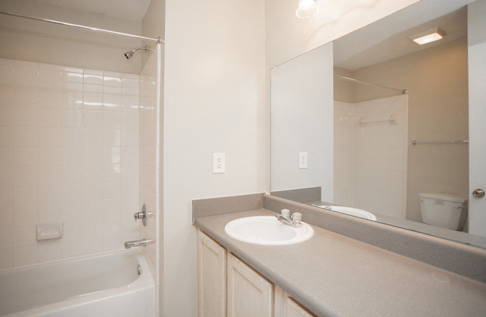 Apartment bathroom featuring a large vanity with a single sink, a wide mirror, and a bathtub with a showerhead and white tile surround. Neutral-colored countertops and cabinetry complement the simple, clean design