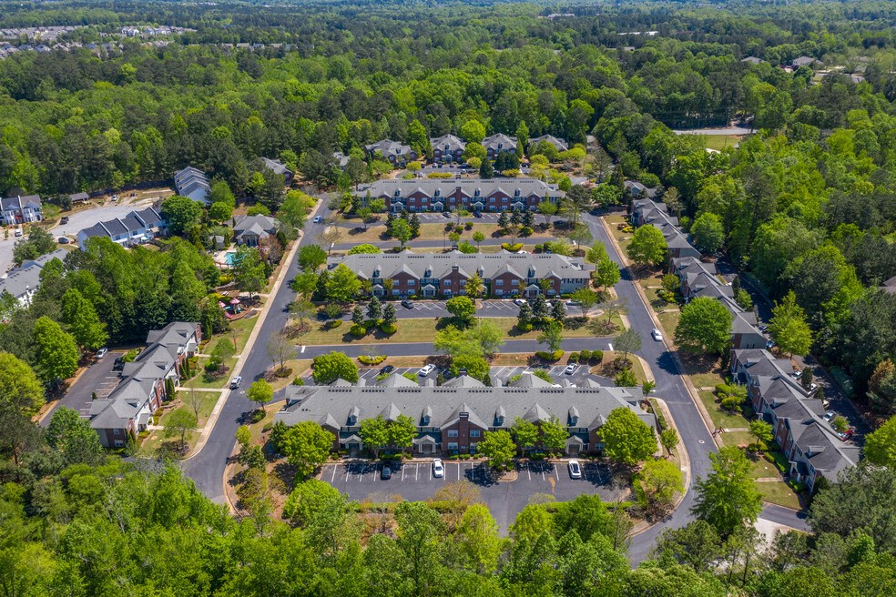 Aerial view of a large apartment community surrounded by lush greenery and wooded areas. The complex features multiple rows of townhome-style buildings with brick exteriors, neatly arranged parking lots, and well-maintained landscaping along winding roads