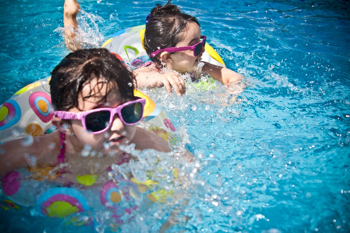 Two children wearing pink sunglasses are swimming in a pool.