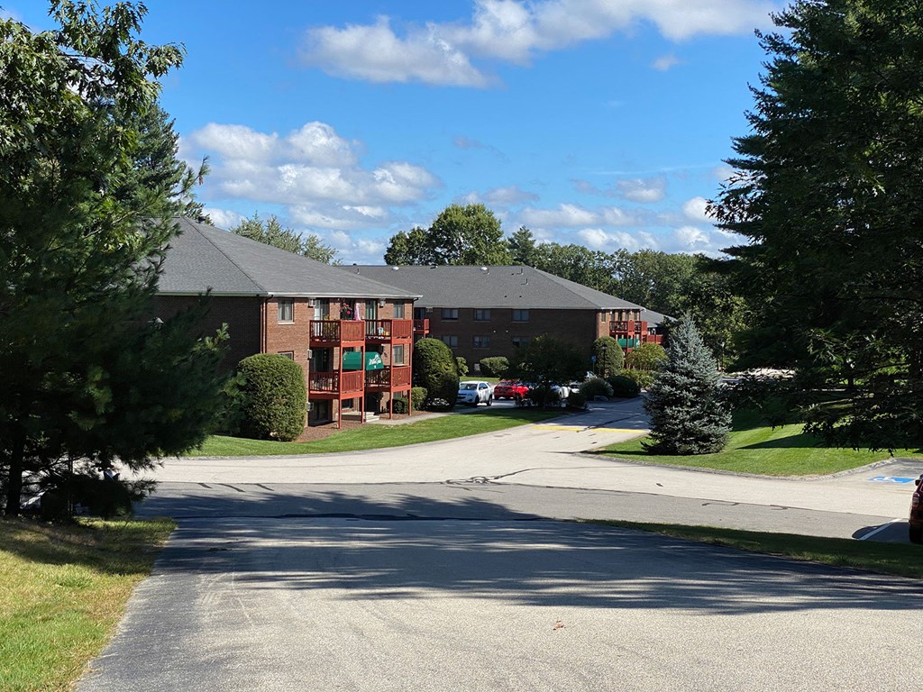 A large red building with a grey roof is surrounded by trees and has a driveway in front.