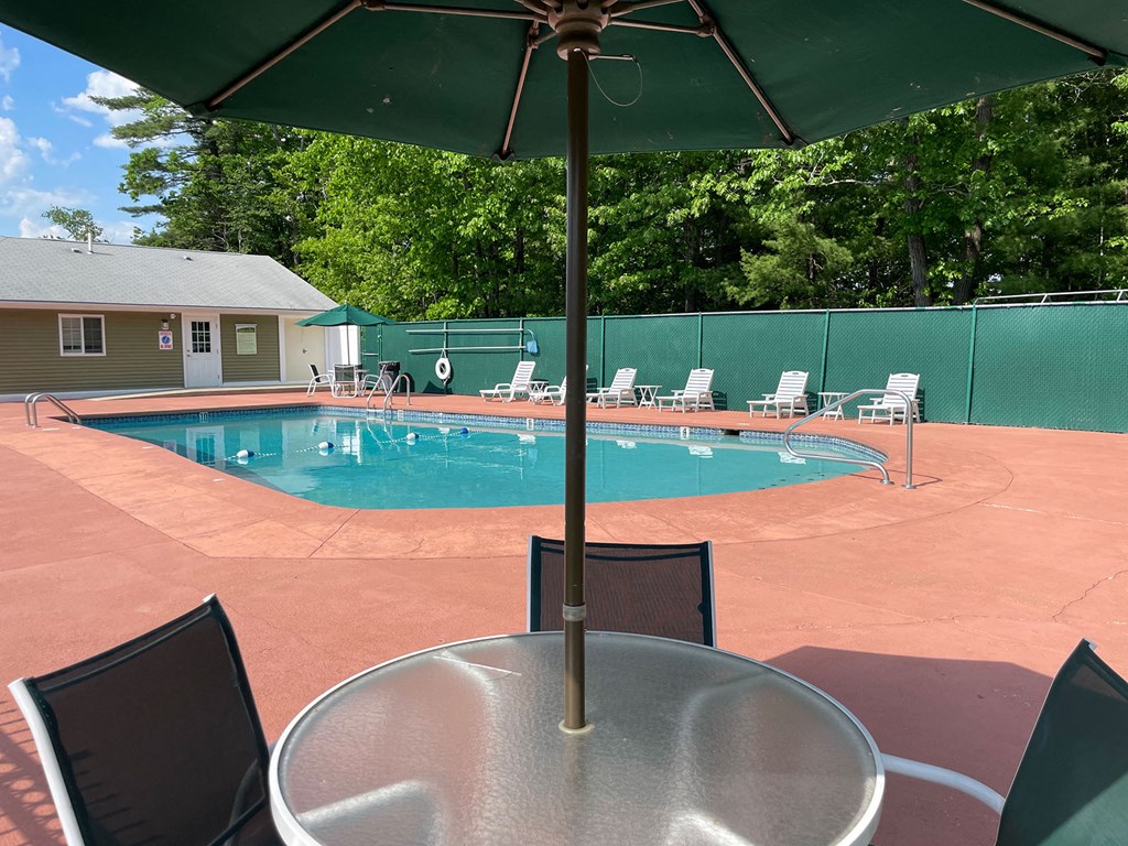 A pool with chairs and umbrellas is surrounded by a green fence.