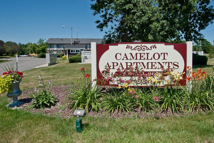 a sign for camlot apartments in front of a yard with flowers