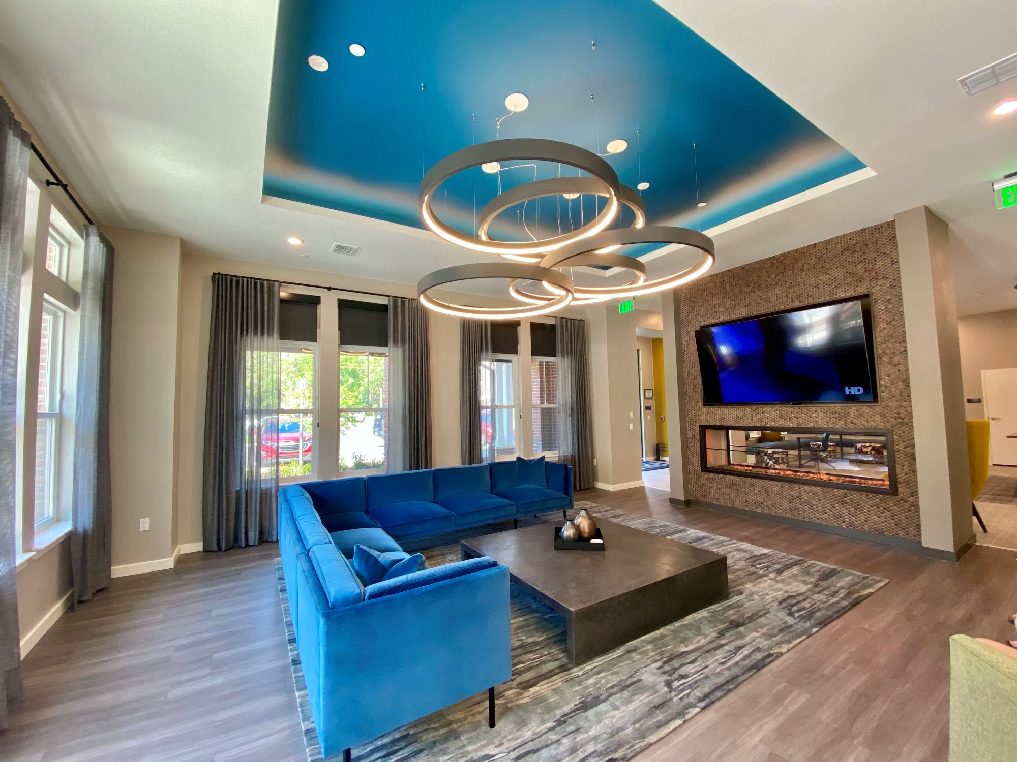 A clubhouse lobby with large windows, a comfortable L-shaped sofa, a large TV, and modern circular chandelier lighting.