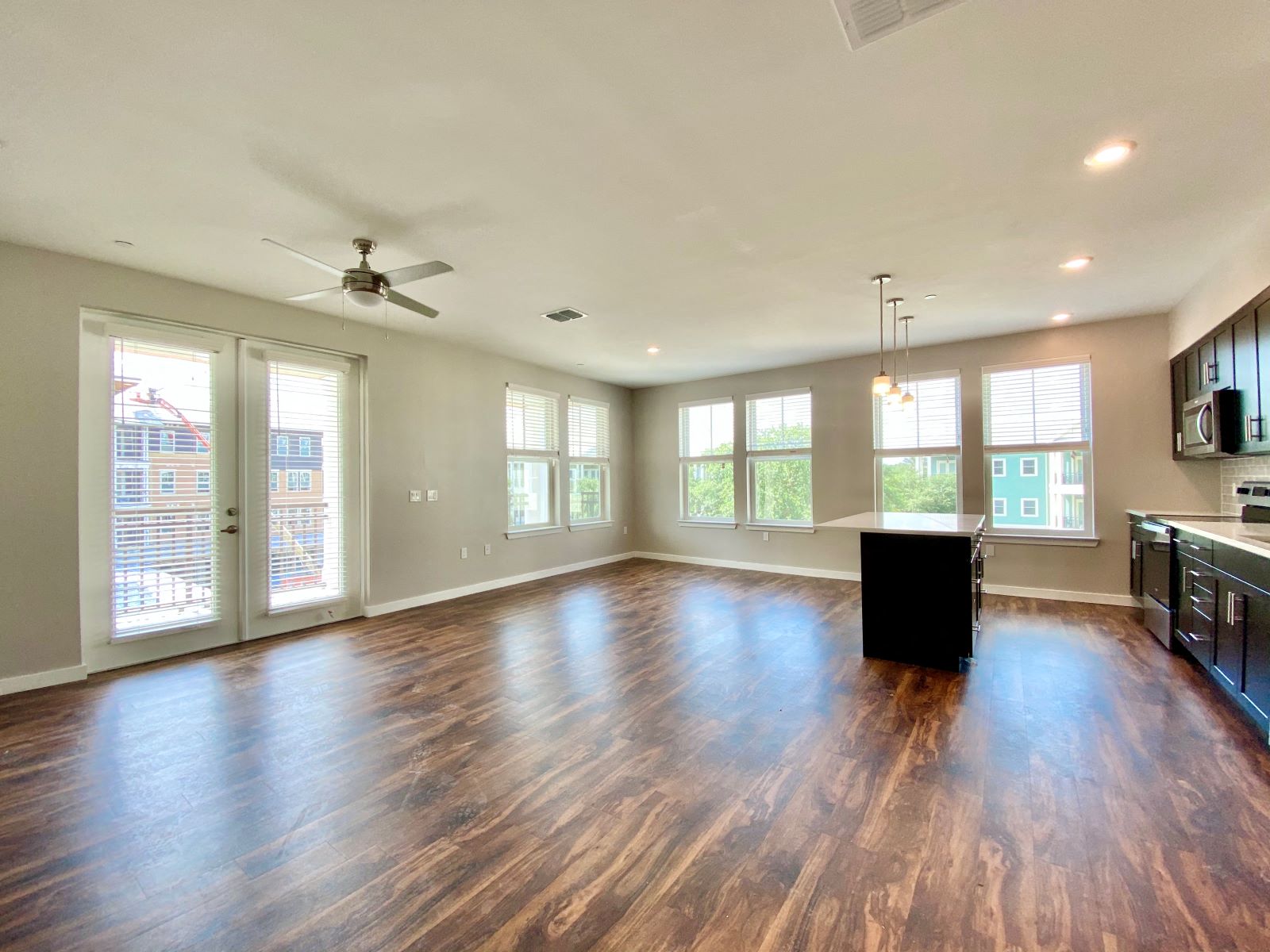 A large open living room with six windows, double doors to a balcony, a ceiling fan, and hardwood-style flooring near a kitchen with an island bar.