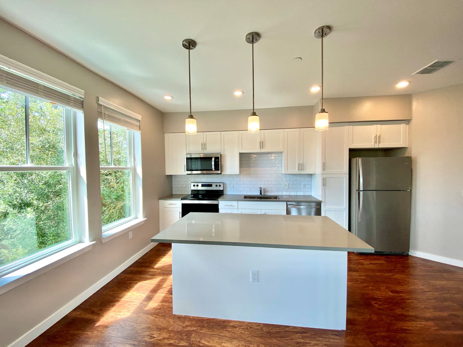 A well-lit open concept kitchen with two large windows, recessed and pendant lighting, hardwood-style flooring, white cabinets, subway tile backsplash, and stainless steel appliances.
