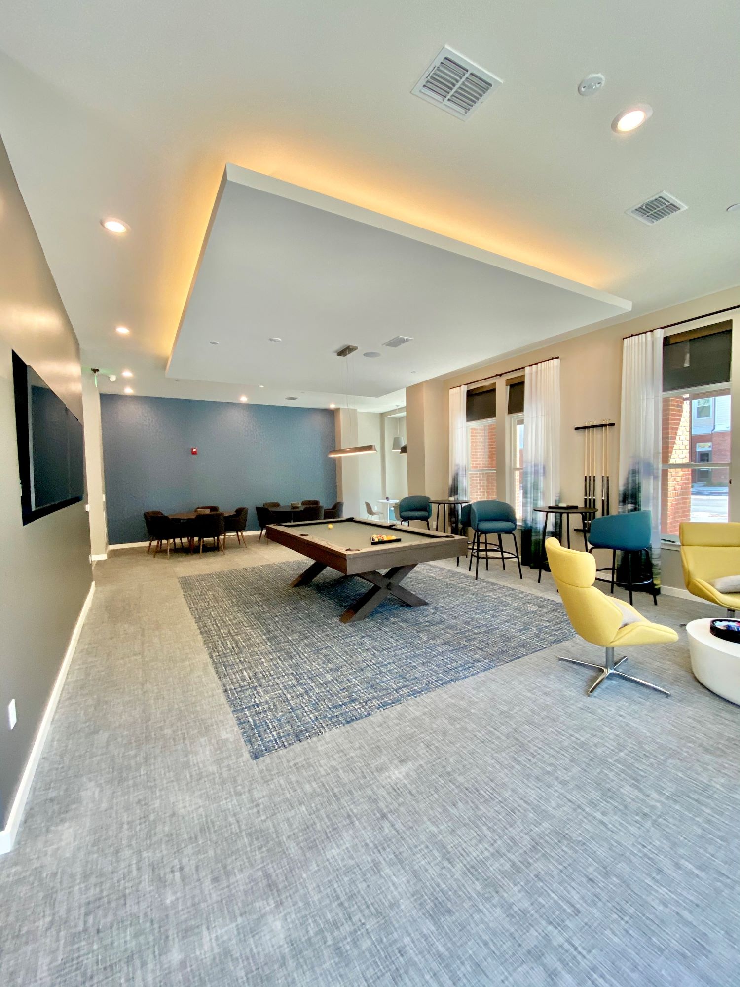 A community game room with several windows, a billiards table and seating areas with chairs and tables.