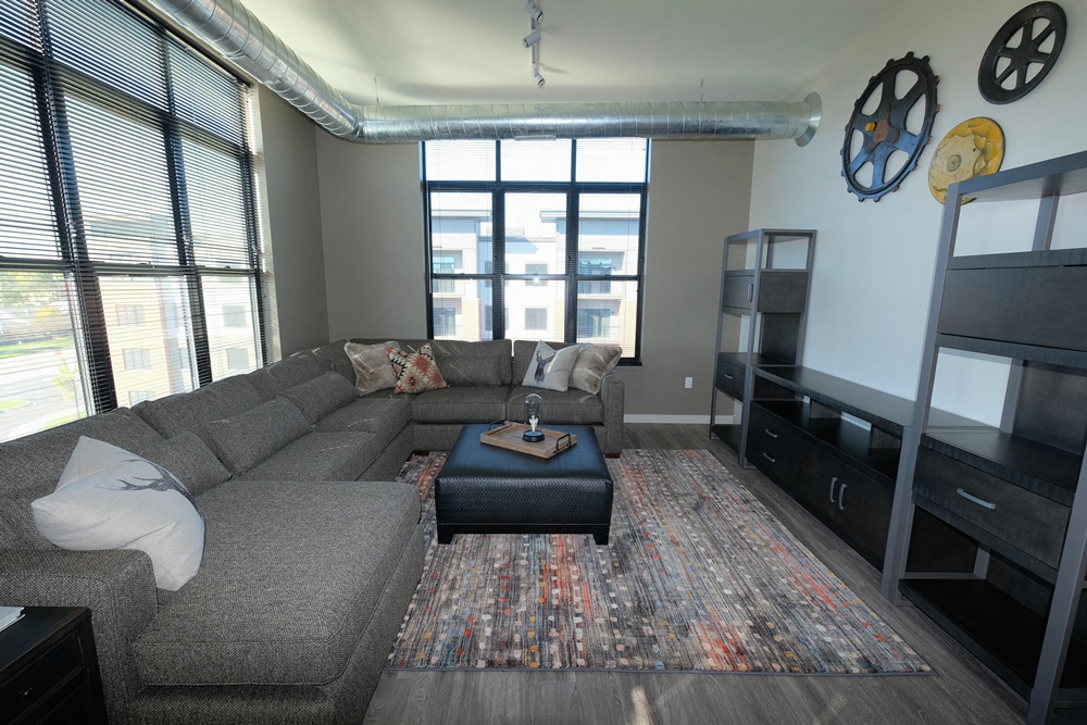 A grey couch with a white pillow sits in a living room with a large window.