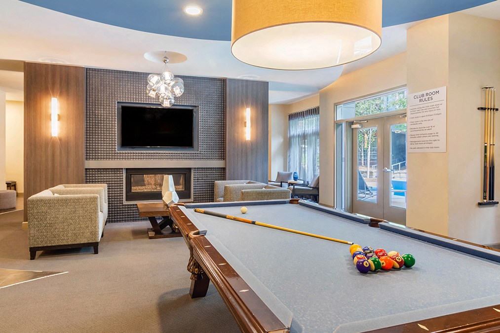 A billiard table with a pool cue and balls is in the foreground of a room with a fireplace and a television.