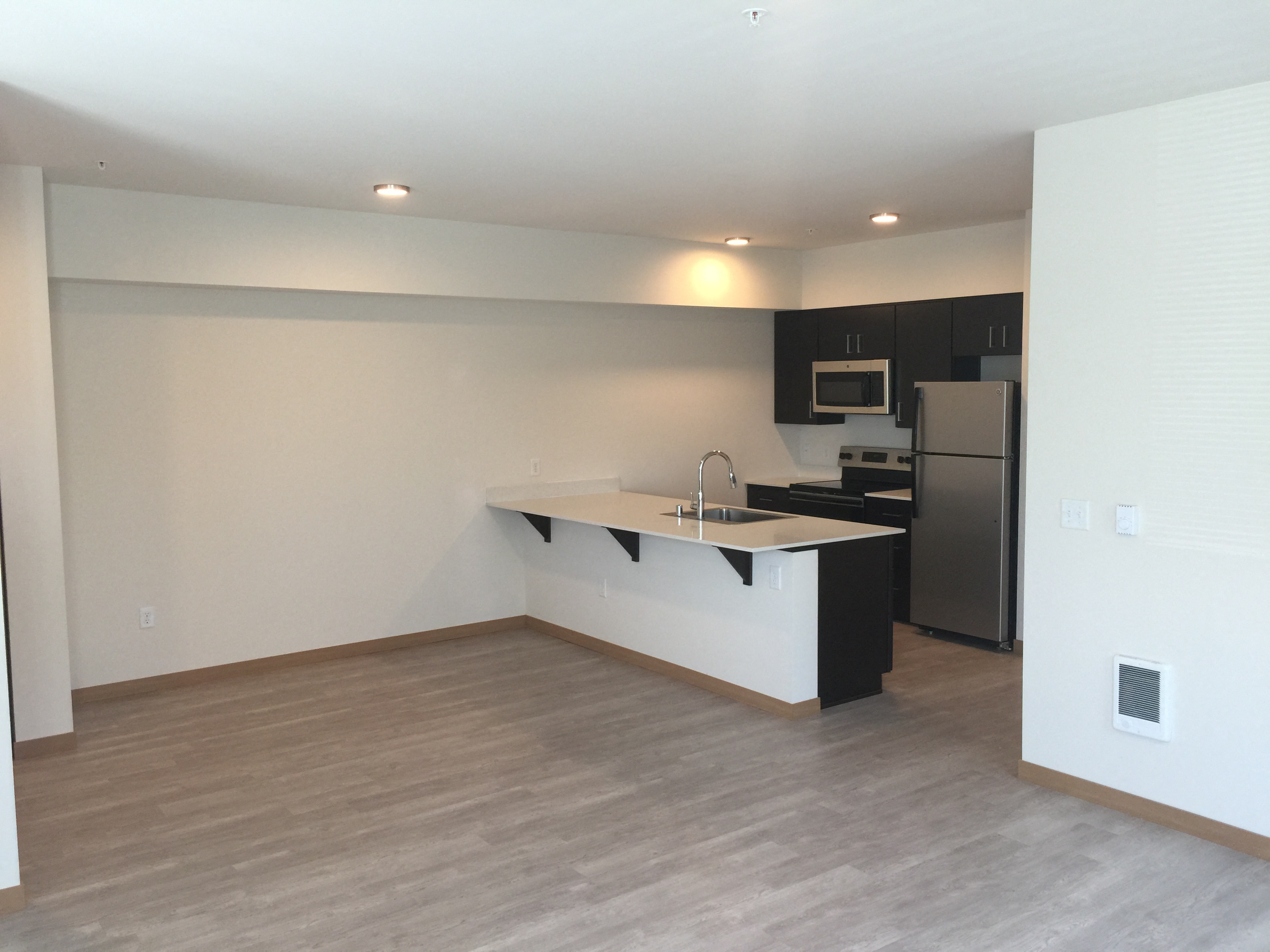 an empty living room with a kitchen and a stainless steel refrigerator