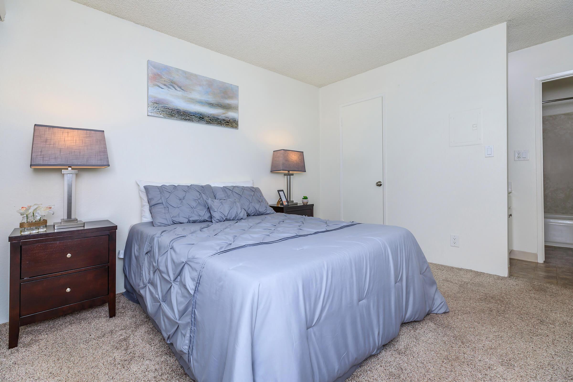 Carpeted apartment bedroom with white walls, bedside tables, and a bed centered against the wall.