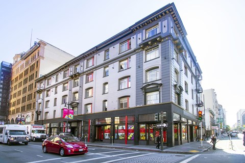 A red car is driving down a street past a building with a black awning.