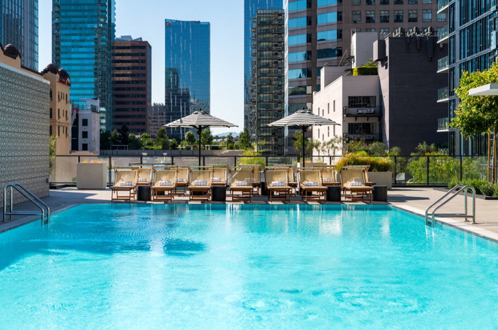 A swimming pool with lounge chairs and umbrellas in the foreground with a city skyline in the background.
