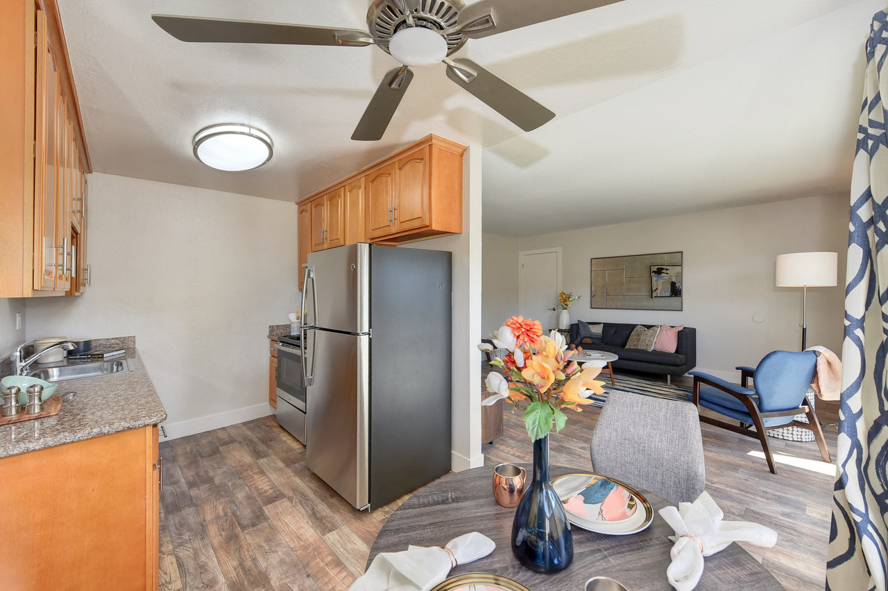 A kitchen with a refrigerator, a ceiling fan, and a dining table with a vase of flowers on it.