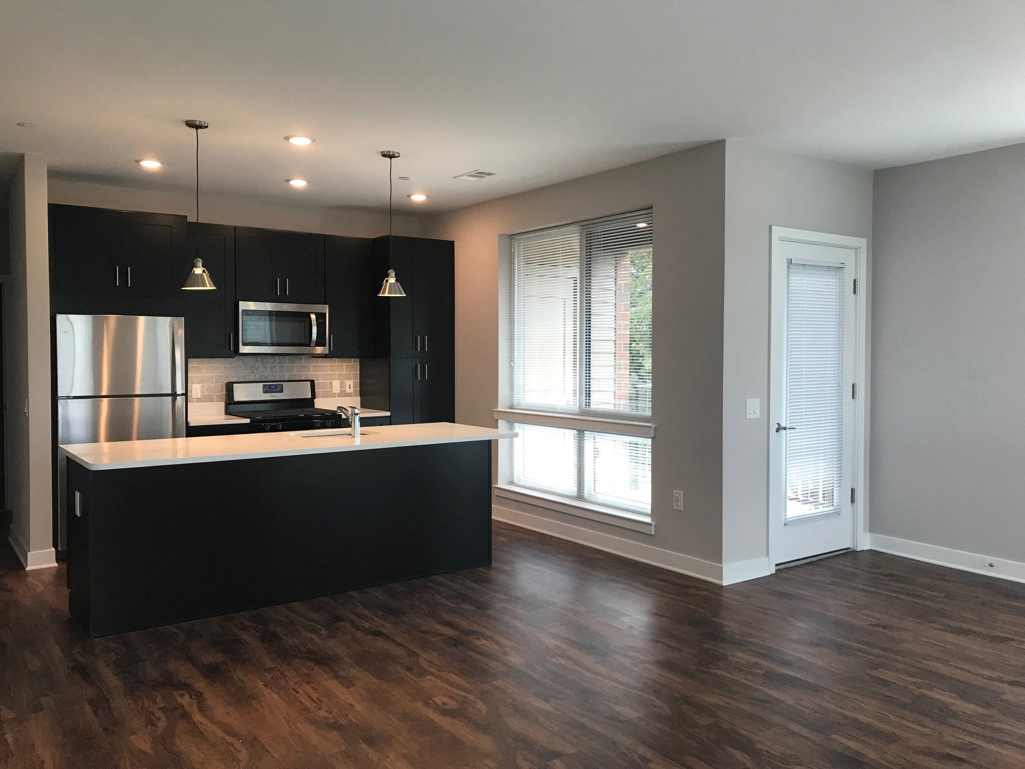 A kitchen with black cabinets, stainless steel appliances, pendant and recessed lighting, a window, and hardwood-style flooring with open access to the living room  at Keva Flats, Exton, 19341.