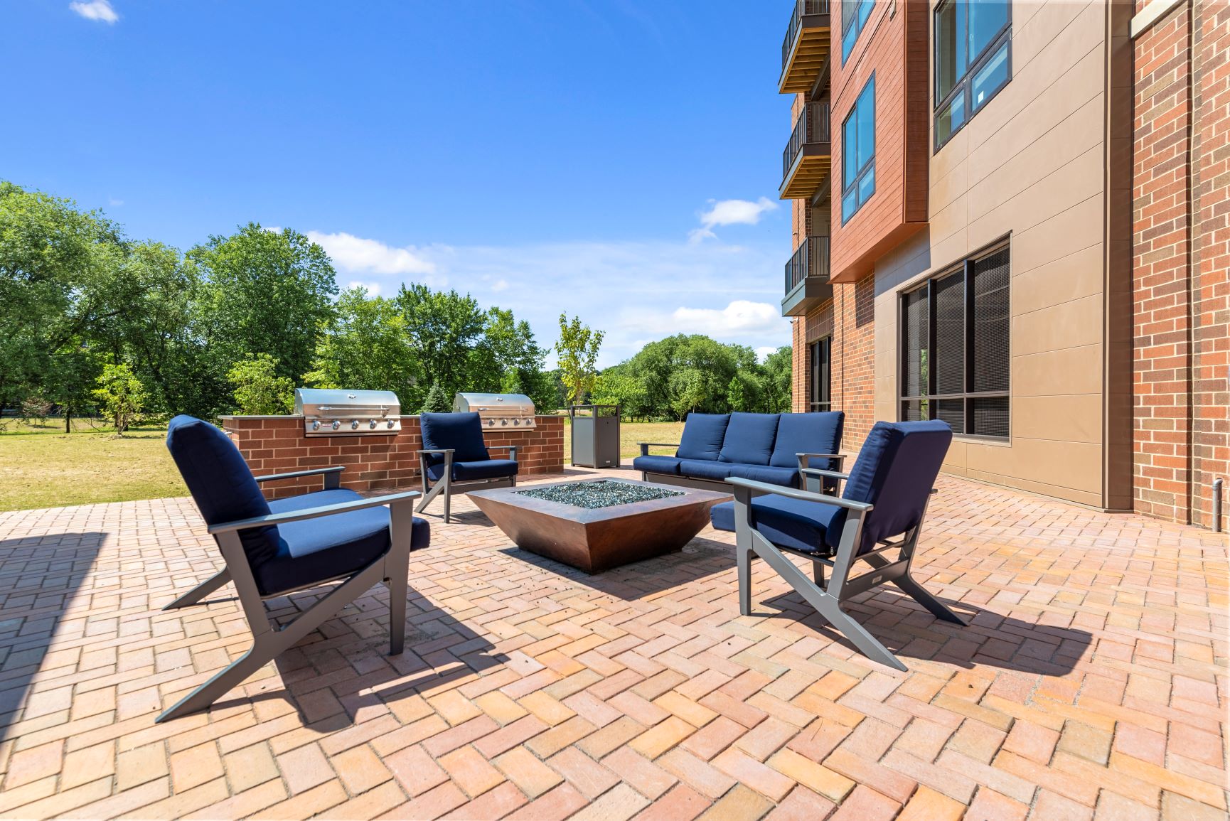An outdoor community patio with a firepit lounge and two barbecue grills near grass and large trees. at Keva Flats, Exton, 19341