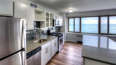 Standing at one end of residence kitchen, the stainless steel appliances and cabinets along the left and a kitchen island on the right. Lake Michigan is seen out the window on the far side of the room
