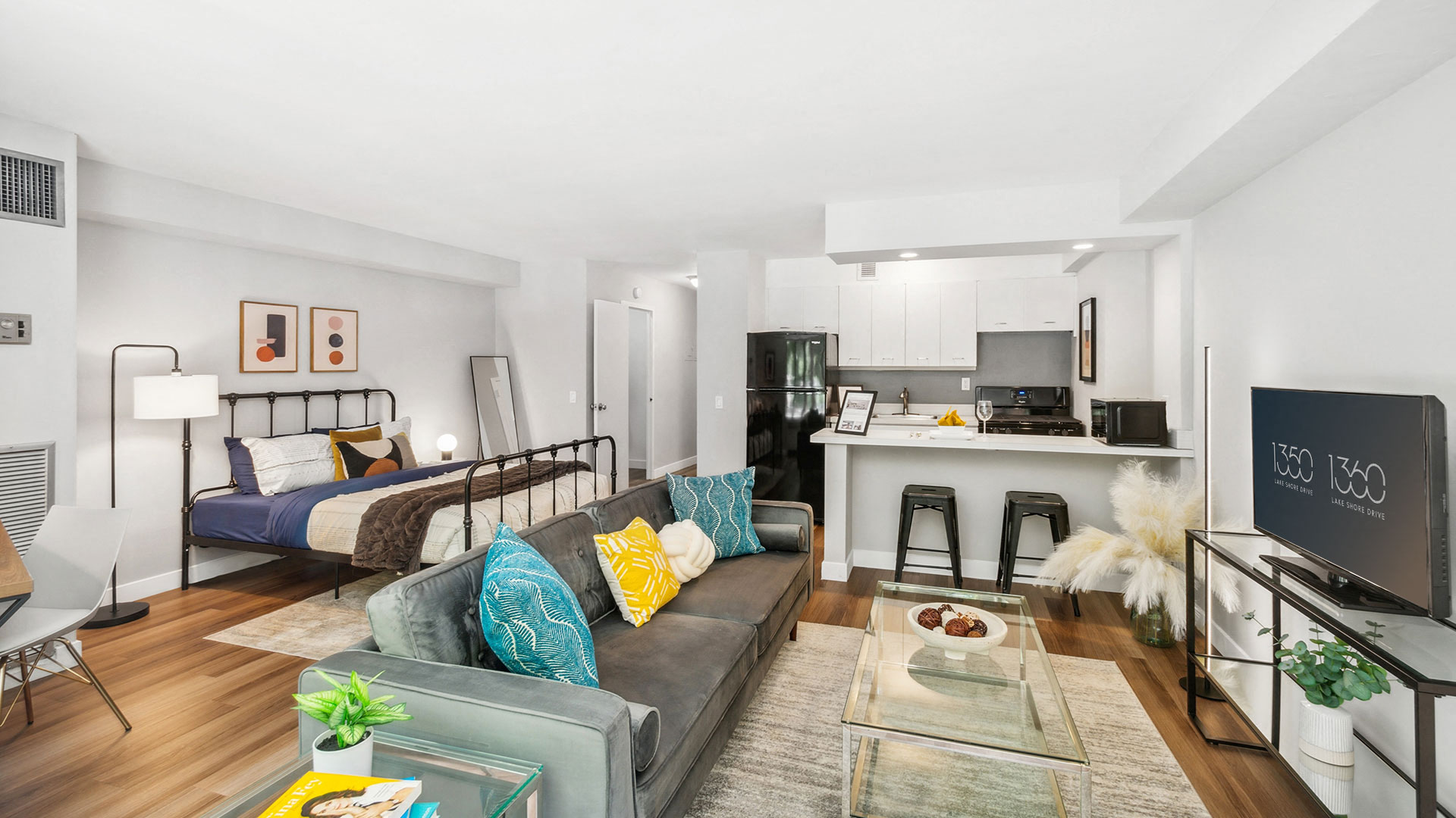 Modern studio apartment with an open layout featuring a bed, a sofa, a TV, a dining table, a kitchen area with barstools, and a bathroom door. Decor includes plants, framed art, and colorful cushions.