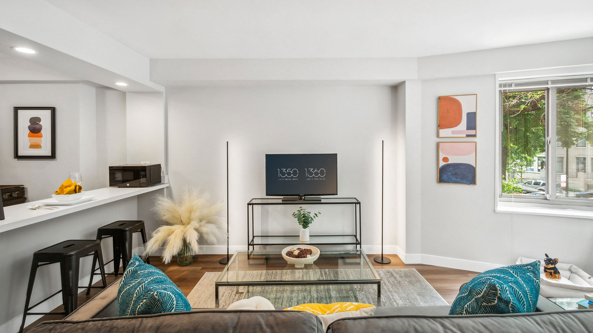 Modern living room with a wall-mounted TV, bar seating area, decorative artwork, and a large window partially covered by blinds.