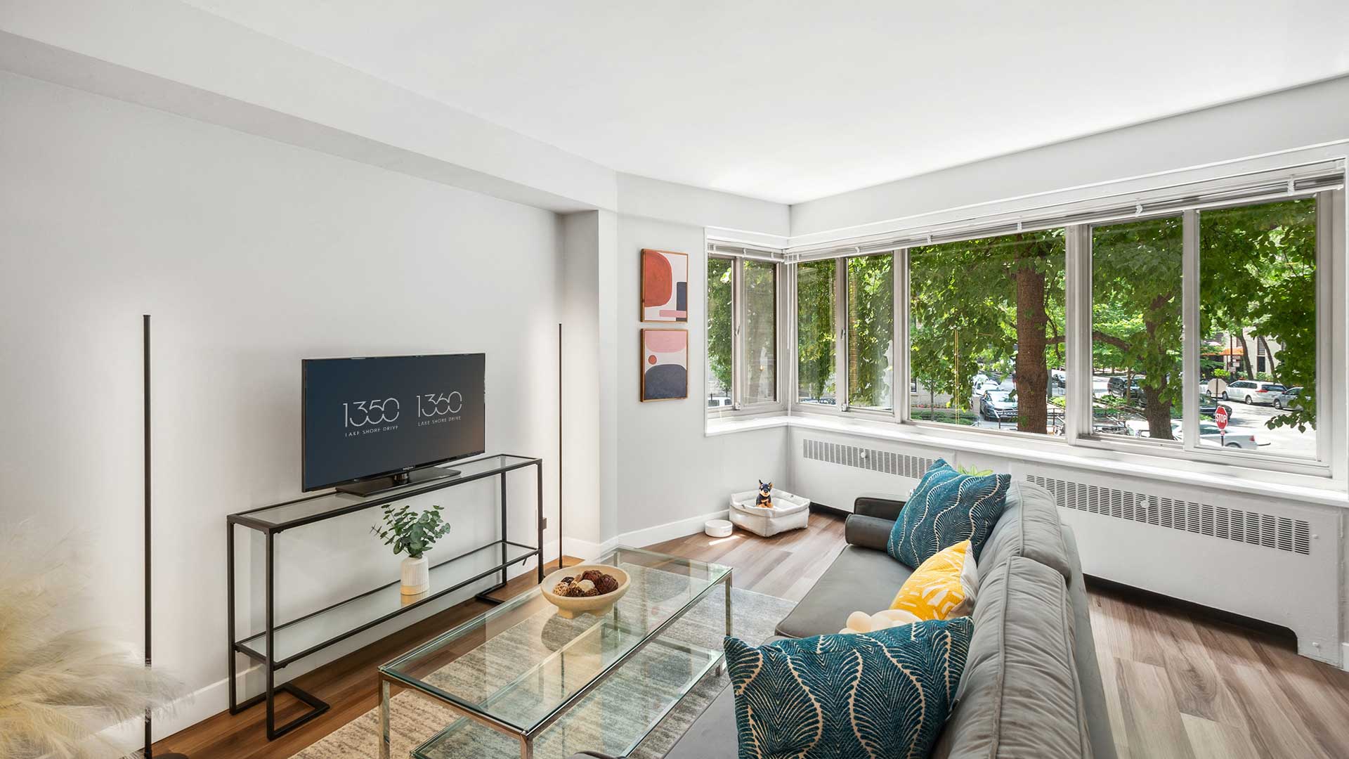 A bright living room with large windows, a grey sectional sofa, a glass coffee table, a wall-mounted TV, and modern decor.