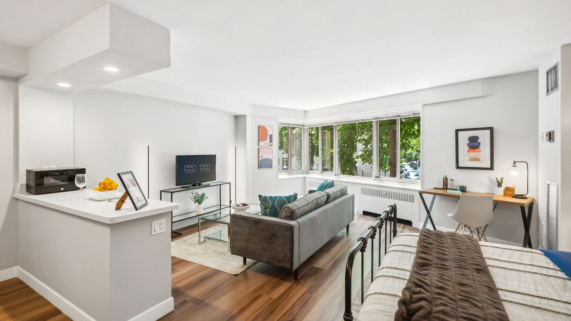 Modern studio apartment with an open floor plan featuring a bed, gray sofa, TV on a stand, a desk with a chair, and a kitchen area. Large window with a view of trees provides natural light.