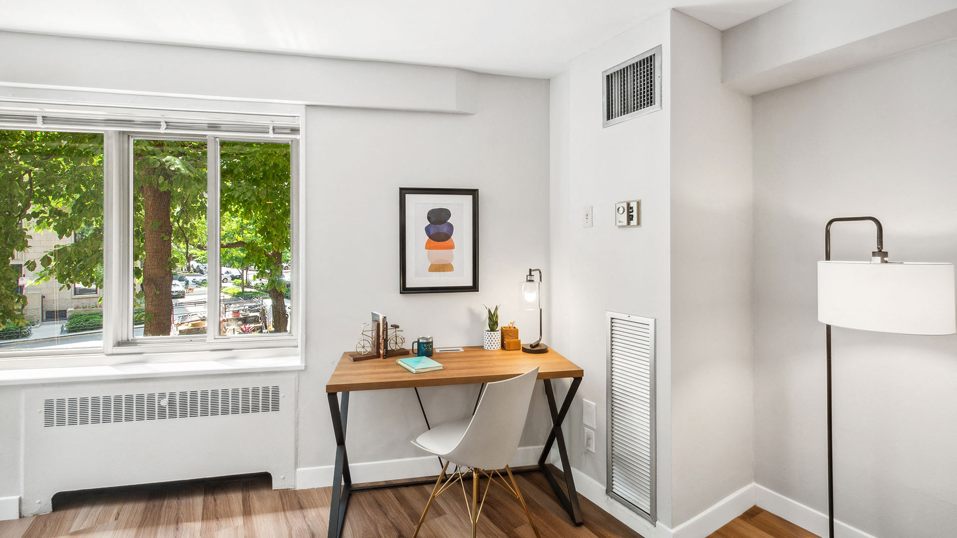 A minimalist workspace with a wooden desk, white chair, and a framed abstract art piece on the wall. A plant, lamp, and books are on the desk. The room has a window with a view of trees outside.