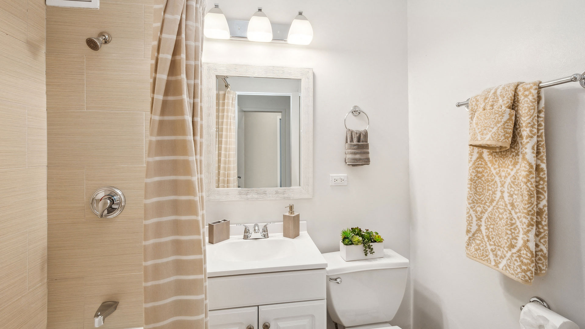 A small bathroom with a white sink, mirror, toilet, shower with a striped curtain, and towel rack holding two patterned towels. A small potted plant sits on the toilet tank.