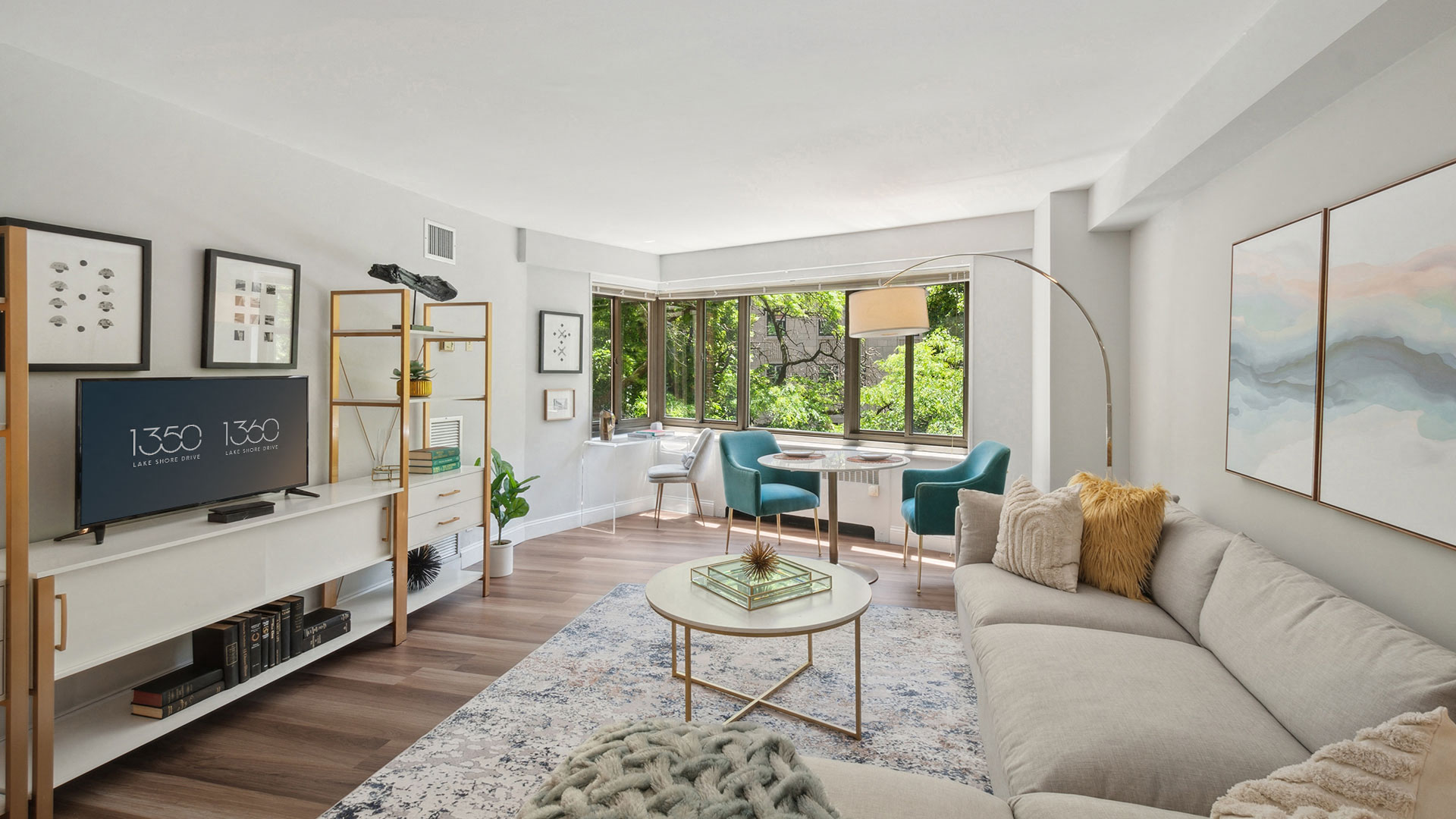 A modern living room with a light beige sectional sofa, round coffee table, television on a white stand, bookshelves, and a small dining area by large windows with green outdoor views.