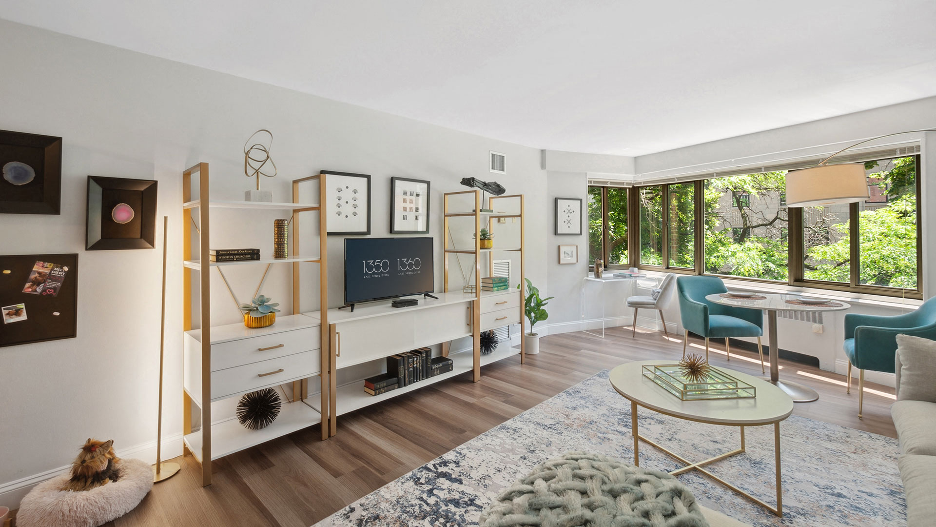 Modern living room with a white media console, bookshelves, and a wall-mounted TV. There are teal chairs by a large window with a garden view, and contemporary decor throughout.