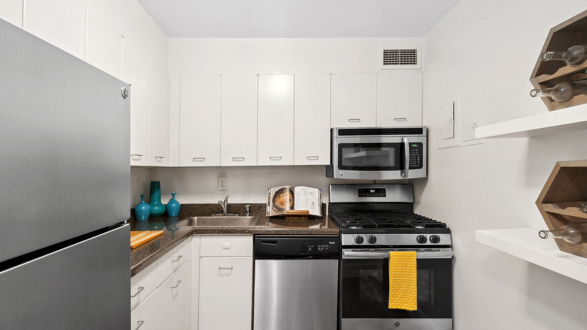 A small kitchen with white cabinets and stainless steel appliances, including a refrigerator, dishwasher, stove, and microwave. There is a cookbook open on a stand near the stove and a yellow towel ha