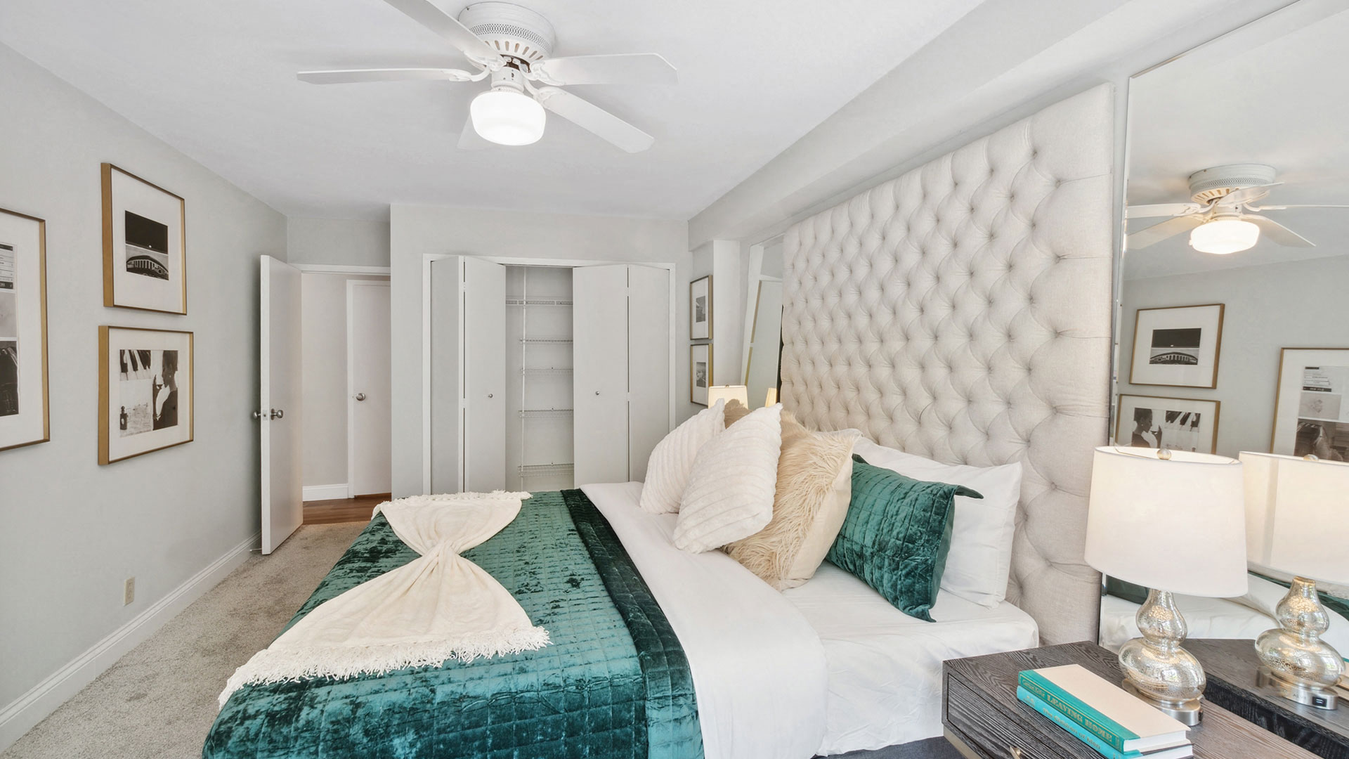 A bedroom features a large tufted headboard, green bedding, a ceiling fan, side table with lamps, and framed artwork on white walls. An open door reveals a closet with shelves.