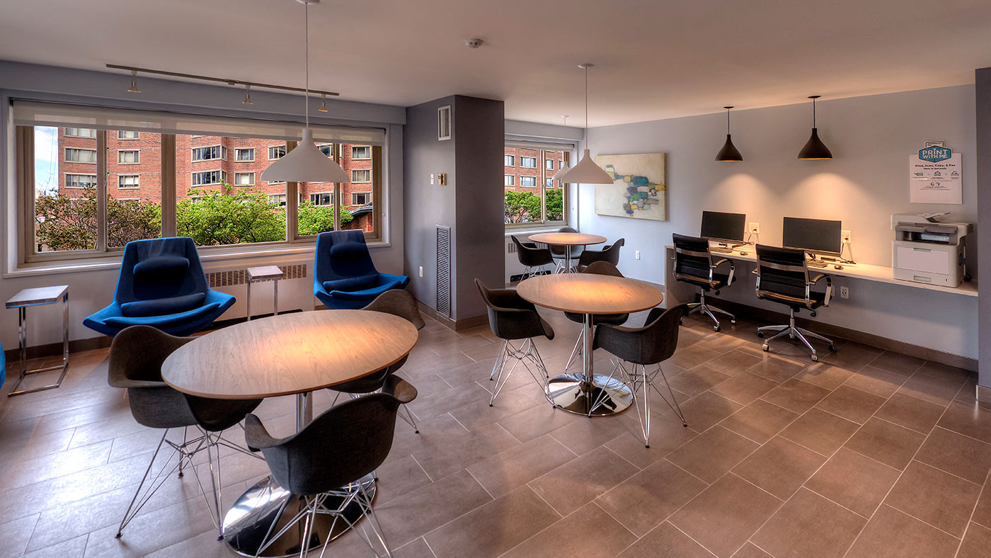 Several round cafe tables are set in the room with club chairs along the far wall below the windows. Along the right wall are two computer stations and a printer.