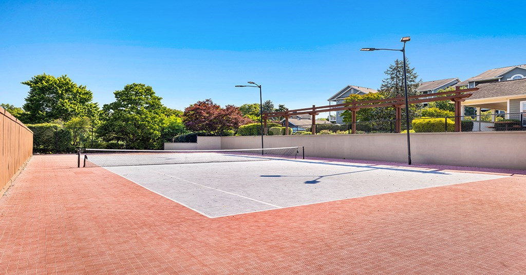 A basketball court with a red and white surface is surrounded by a fence and houses.