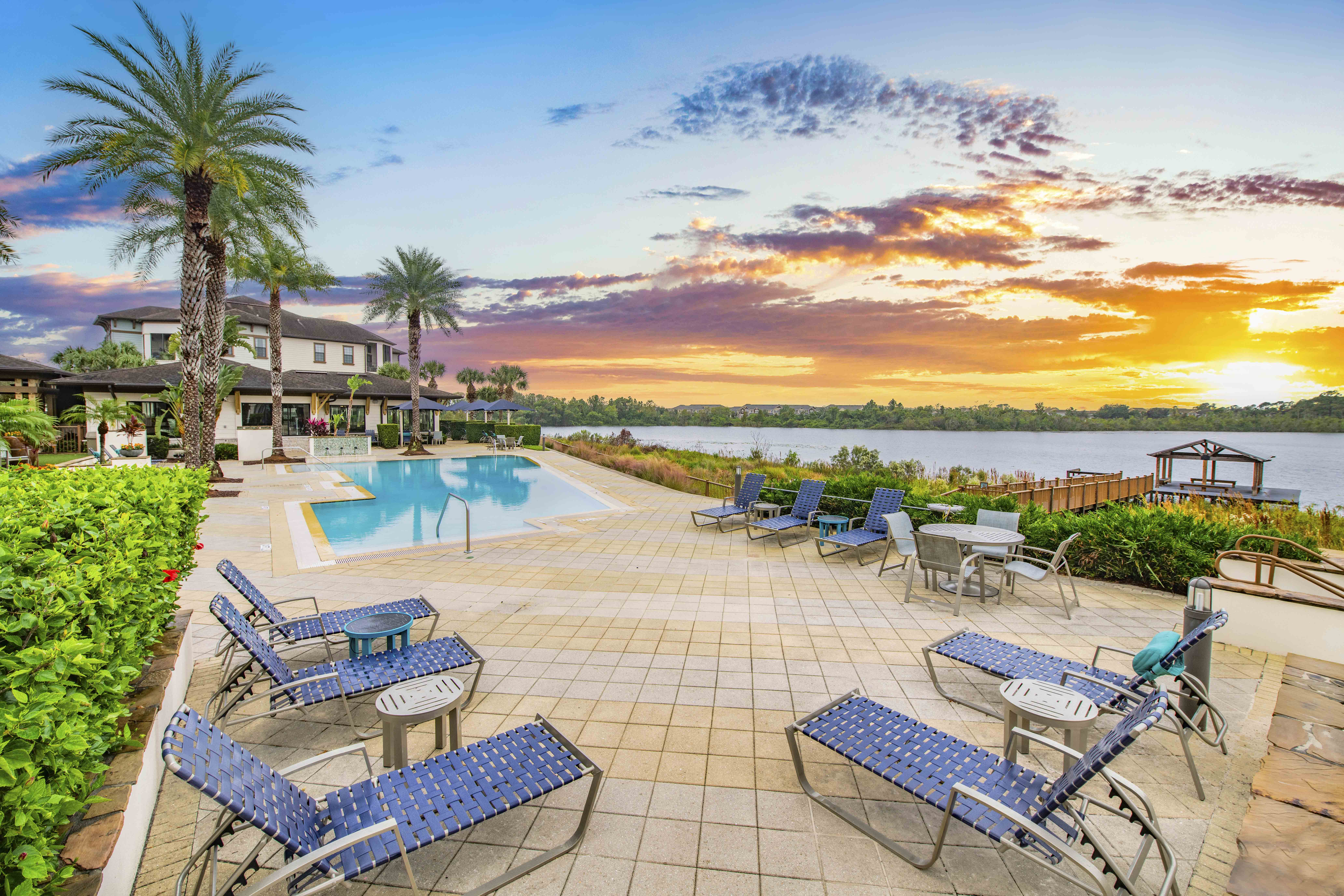a pool with blue chairs and a poolside patio with a sunset in the background