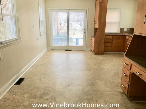 A room with a sliding glass door leading to a balcony, a window, and a kitchen area with wooden cabinets and a countertop.