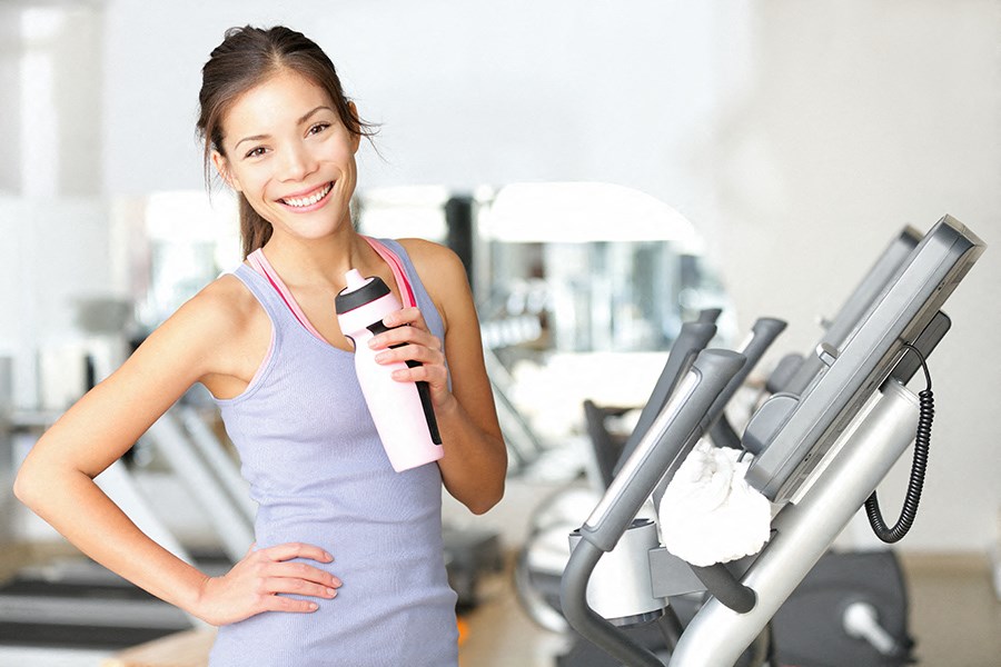 a woman holding a bottle of water and standing next to a gym treadmill