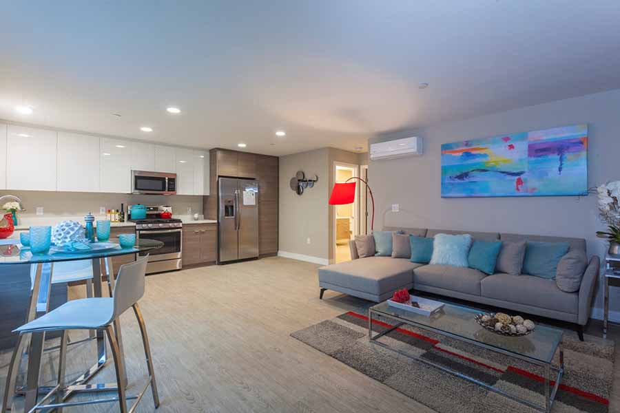 Pet Friendly Apartments in Campbell CA - Parc at Pruneyard - Living Area with L-Shaped Couch, Glass Coffee Table, Rug, Wood Flooring, Brightly-Colored Framed Wall Art, with View of Dining Table with High-Top Seating, and the Kitchen