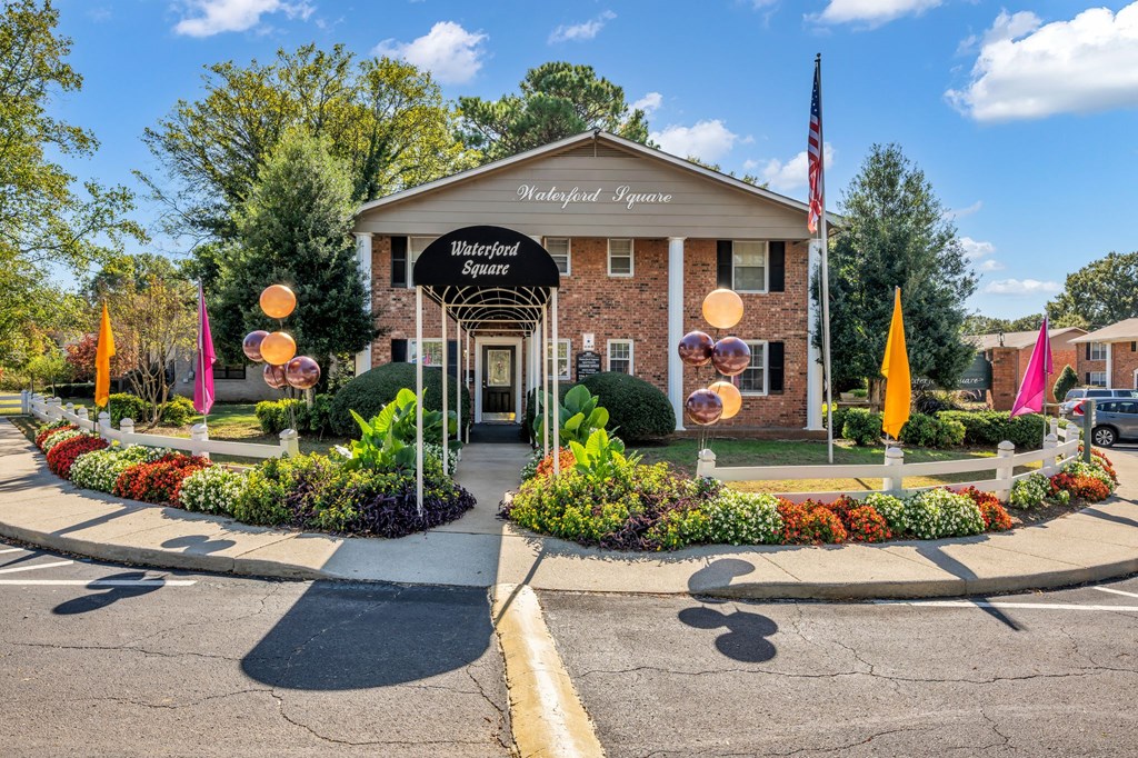 The image shows a building with the name "Waterford Square" on it, surrounded by a colorful flower bed and a clear blue sky.
