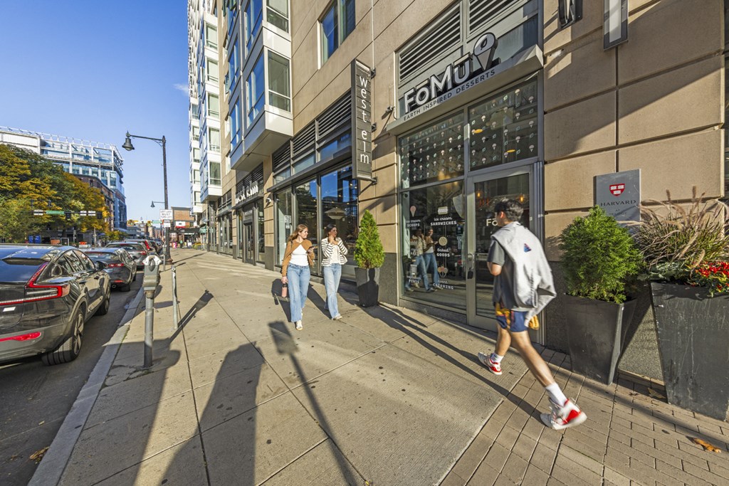 A man in a grey hoodie and blue shorts is walking down a sidewalk past a store with a sign that reads "FORMULA".