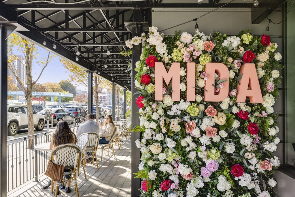 People are sitting at tables under a canopy with a flower wall spelling out MIDA.