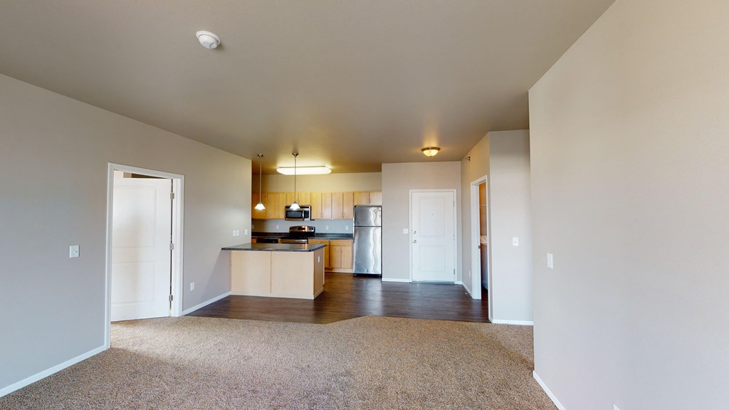 an empty living room with a kitchen with a refrigerator and a sink
