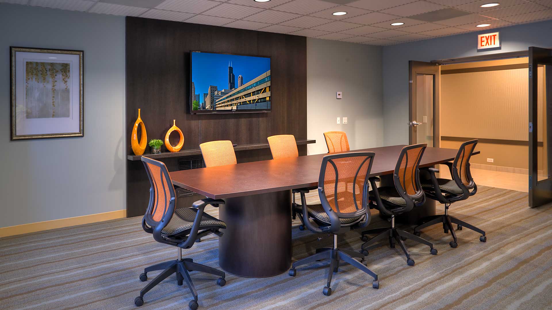 A conference room table and chairs in the conference room. A television is mounted on the left wall and the doors are open to the right.